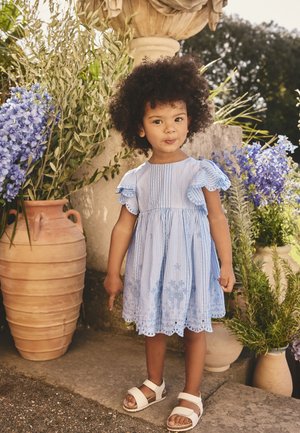 Jeune fille en robe rayée bleue et sandales blanches se tient à l'extérieur près de grands pots en terre cuite avec des fleurs bleues et de la verdure.