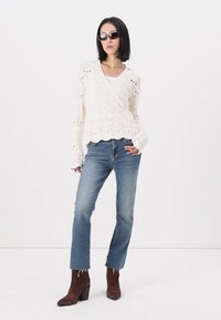 Cremfarbener Häkelpullover mit gewelltem Saum, kombiniert mit verwaschenen blauen Jeans und dunkelbraunen Knöchelstiefeln. Model trägt dunkle Sonnenbrillen.