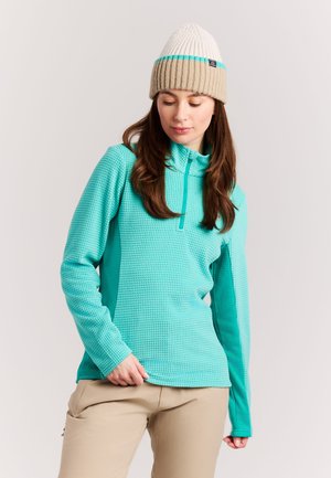 Vrouw draagt een beige-witte gebreide muts en een turquoise trui met lange mouwen en beige broek, kijkt naar beneden.