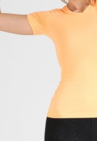 Vaaleanoranssi urheilupaita, jossa on lyhyet hihat, slim fit ja sileä rakenne; yhdistettynä mustiin leggings-housuihin, joissa on hienovarainen kuvio.