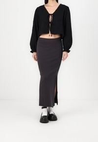 Blouse noire courte à manches longues avec un détail noué sur l'avant, associée à une jupe crayon grise fendue sur le côté et des bottines.