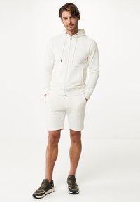 Vitt zip-up hoodie med dragsko, i kombination med vita shorts. Bärs med mörka sneakers med ljusare accenter, mot en enkel bakgrund.