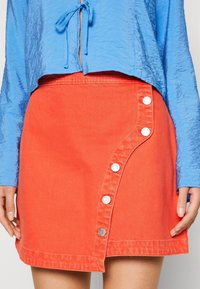 Jupe en denim corail avec une patte de boutonnage diagonale comportant cinq boutons blancs ; complétée par un crop top bleu texturé.