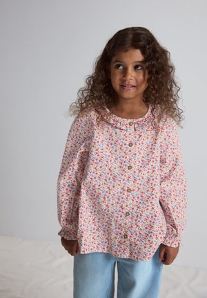 Jeune fille aux cheveux bouclés portant un chemisier à manches longues à motifs floraux et un jean bleu clair, regardant légèrement vers sa droite.