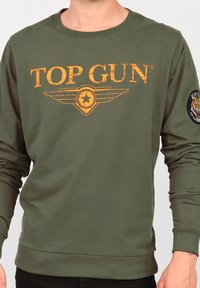 Olivgrön sweatshirt med "TOP GUN"-logotyp i sliten gul färg, med en stjärna och vinggrafik; vänster ärm har en rund militärstilstämpel.