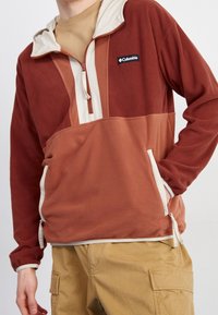 Pullover in pile color-blocked nelle tonalità di ruggine e beige, con chiusura a mezza zip, cappuccio regolabile e tasche laterali con bordo a contrasto.