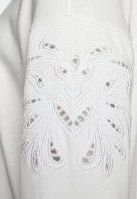 Felpa bianca con intricati ricami a forma di fiori, che dettagliate forme a goccia e un design a cuore. Texture morbida.