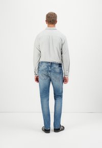 Camicia grigia chiaro a maniche lunghe, jeans in denim blu e scarpe nere, visti da dietro, che mostrano cuciture e tasche posteriori.