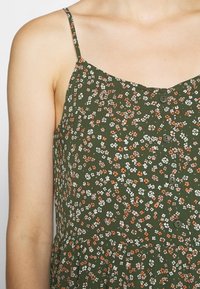 Gros plan d’une robe verte à fines bretelles, décorée de petits motifs floraux blancs, orange et rose et de quatre boutons foncés sur le devant.