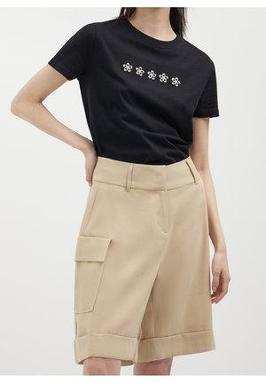 Kvinde iført en sort t-shirt med fem blomsterpynt og beige knælange cargoshorts med opkrogede kanter og sidelommer.