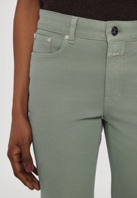 Gros plan sur un pantalon vert clair avec des poches avant et une braguette à boutons porté par une personne à la peau foncée et aux ongles peints.