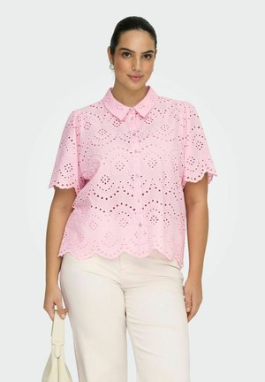 Mujer de cabello oscuro con blusa rosa de encaje con ojales y pantalones color crema, sosteniendo un pequeño bolso blanco sobre un fondo liso.