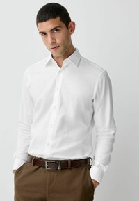 Weißes Langarmhemd mit geknöpfter Front, spitzem Kragen und glatter Textur, kombiniert mit braunen Hosen und einem braunen Gürtel.