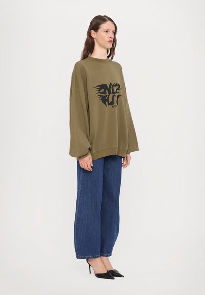 Olivenfarvet oversized sweatshirt med sort grafisk design på forsiden, parret med brede blå denimjeans og spidse sorte hæle.