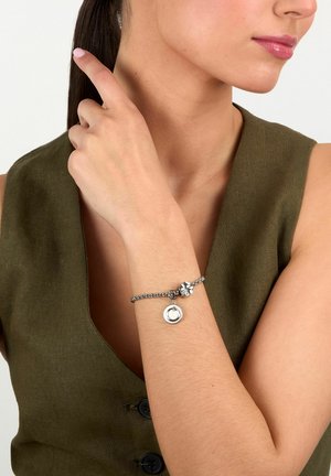 Donna con top verde oliva senza maniche indica la coda di cavallo mostrando un bracciale d'argento con ciondoli rotondi e a forma di fiore al polso.