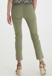 Pantalons ajustés verts avec une texture lisse, longueur cropped et ourlet roulé, associés à des sandales compensées beiges. Vue arrière montrée.