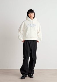 White fleece hoodie with front pocket and blue "tommy" logo, paired with loose black pants and black shoes on a neutral background.