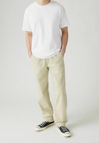 Personne portant un t-shirt blanc uni, un pantalon beige avec les mains dans les poches, et des baskets basses noires avec des semelles et des lacets blancs.