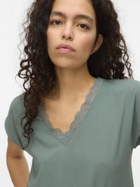 Young woman with curly dark hair wearing a green V-neck top with lace trim, looking directly at the camera against a plain background.