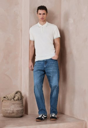 Bărbat stând lângă un perete bej, purtând un polo alb cu mâneci scurte, blugi albaștri și adidași alb-negru, cu mâna în buzunar.