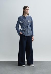Γυναίκα με μπλε υφασμάτινο σακάκι με ζώνη και τσέπες με λευκή επένδυση, και φαρδιά σκούρα μπλε παντελόνια, όρθια με το ένα χέρι στην τσέπη.