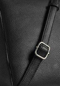 Borsa in pelle nera con texture in primo piano che mostra una tracolla regolabile con una fibbia quadrata in argento.