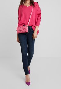 Suéter rosa de cuello en V, jeans skinny oscuros y tacones altos morados. El bolso cruzado rosa presenta un patrón de formas azules.