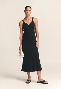 Črna brezrokavna obleka z V-izrezom, naborki na prsih in lahkotnim robom. Kombinirana s črnimi sandali in bež teksturirano torbico.