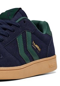 Marinmockad sneaker i mocka med gröna läderdetaljer, med strukturerad gummisula och guld "hummel"-logotyp på sidan.