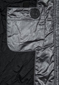 Interno di una giacca nera trapuntata con una tasca grigia a scacchi dotata di una grande chiusura a bottone nero. Materiale liscio e lucido.