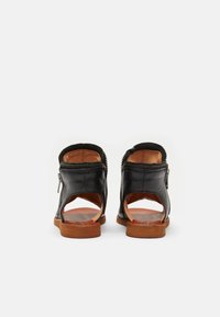 Sandalias de cuero negro con parte trasera abierta, diseño angular, cierre con cremallera y suela de goma marrón. Textura suave y detalles mínimos.