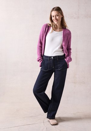 Cardigan en tricot rose, top blanc, et jeans larges bleu marine. Boutons roses sur le cardigan, couture subtile sur les jeans, et chaussures beiges.