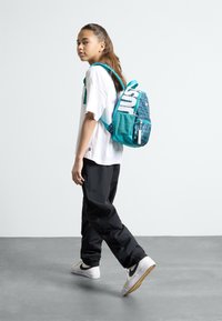 Τσάντα πλάτης σε τιρκουάζ χρώμα με γεωμετρικά σχέδια, μπροστινός θήκη με φερμουάρ και λευκό κείμενο "ΑΠΛΑ ΚΑΝΕ ΤΟ". Το παιδί φοράει λευκό μπλουζάκι και μαύρο παντελόνι.