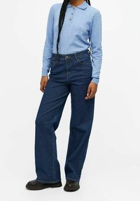 Personne portant un polo à manches longues bleu clair, un jean bleu foncé taille haute à jambes larges et des chaussures noires, debout devant un fond blanc.