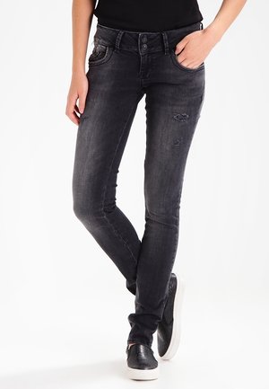 Kvinde iført tætsiddende sorte slidt jeans og sorte slip-on sko, stående med den ene hånd i lommen mod hvid baggrund.