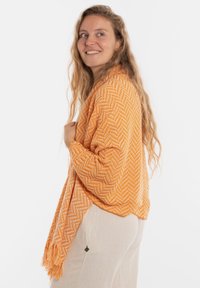 Oranje geblokte cardigan met een visgraatpatroon, voorzien van franjes aan de randen en een losse pasvorm, gedragen over lichtgekleurde broeken.
