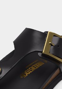 Μαύρο λουράκι δερμάτινου σανδαλιού με χρυσή πόρπη και ορατό το όνομα του εμπορικού σήματος "Birkenstock" στο πέλμα.