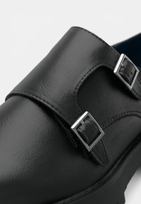 Μαύρο δερμάτινο παπούτσι slip-on με λεία υφή, διπλό μεταλλικό κούμπωμα και στρογγυλεμένη μύτη με ραφές σε αντίθεση.