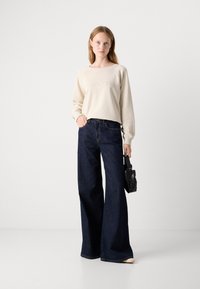 Felpa crema con testo in rilievo, abbinata a jeans a gamba larga blu scuro. Una piccola borsa nera completa l'outfit. Presentazione pulita e minimalista.