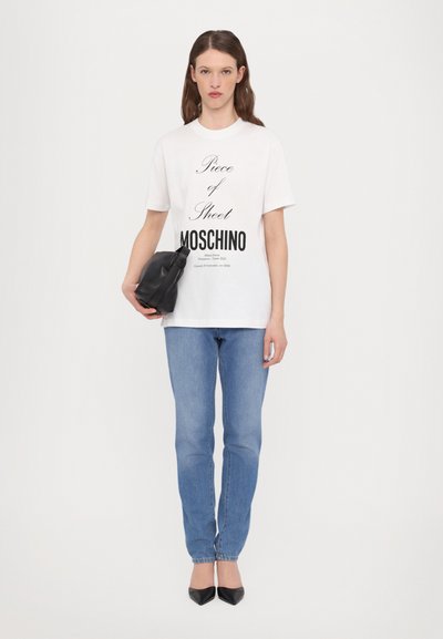 White cotton t-shirt with black printed text, paired with light blue fitted jeans and black pointed-toe heels; holding a black clutch bag.