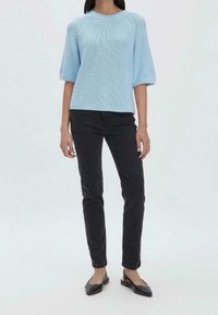 Femme portant un pull en tricot bleu clair, un pantalon noir slim et des ballerines à bride arrière noires à bout pointu, debout devant un fond uni.