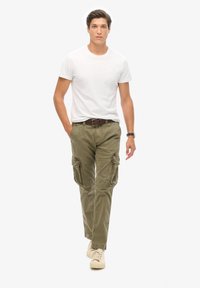 Pantalones cargo verde oliva con múltiples bolsillos laterales, combinados con una camiseta blanca de manga corta y zapatillas beige. Modelo caminando hacia adelante.