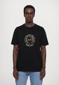 T-shirt en coton noir avec un logo rond doré portant le texte « A|X » et « ARMANI EXCHANGE », manches courtes et coupe décontractée.