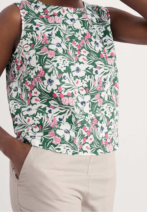 Sleeveless green top with white and pink floral pattern worn with light beige pants, hand in pocket, arm raised.