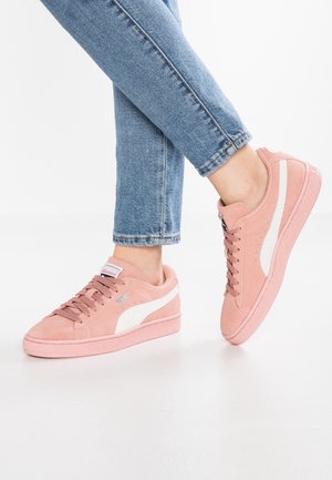 Osoba w jasnoniebieskich dżinsach i różowych zamszowych butach Puma z białymi paskami, stojąca z jedną lekko uniesioną stopą.