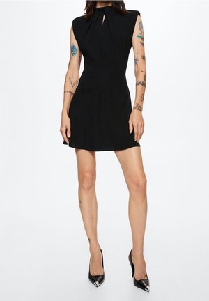 Robe noire sans manches avec un bustier ajusté, détails plissés au niveau du col, jupe évasée et tissu lisse. Associée à des escarpins noirs à bout pointu.