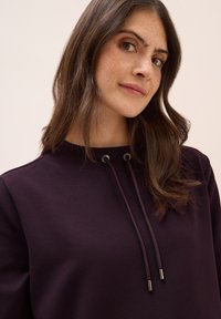 Donkerpaars hoodie met een hoge hals, voorzien van een trekkoord en metalen oogjes. Gelaagd stof en een relaxte pasvorm.