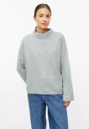 Vrouw draagt een losse lichtgrijze trui met een hoge ronde hals en blauwe spijkerbroek, staand tegen een effen witte achtergrond.