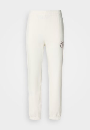 Pantalones de chándal blancos con cinturilla elástica y puños en los tobillos. Presenta un pequeño logo marrón en el muslo izquierdo. Textura de tela suave y lisa.