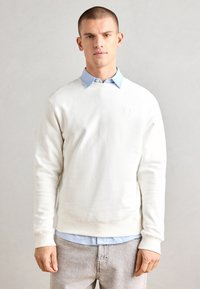 White crewneck sweatshirt made of cotton blend, featuring ribbed cuffs and hem. A light blue collared shirt is layered underneath.